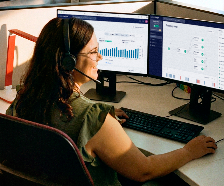 A woman wearing glasses and a professional headset works at a dual-monitor workstation displaying a network monitoring dashboard with bar charts on the left screen and a Topology Map with connected nodes on the right screen. The desk setup includes a keyboard and mouse on a white surface in an office cubicle environment.